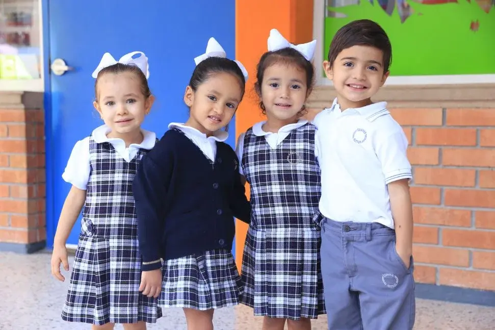 Cuatro estudiantes de primaria en uniforme escolar con moños blancos posando juntos en el colegio.